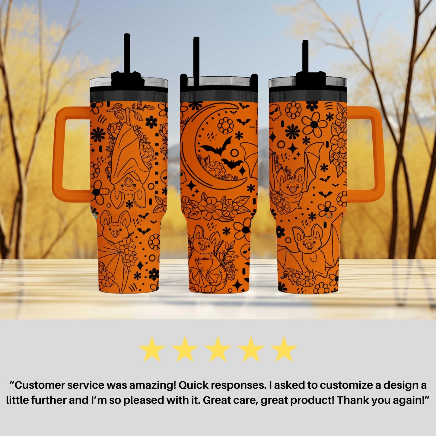 three orange tumblers with black designs, including bats, flowers, and a crescent moon, placed on a wooden surface against a blurred background of trees.