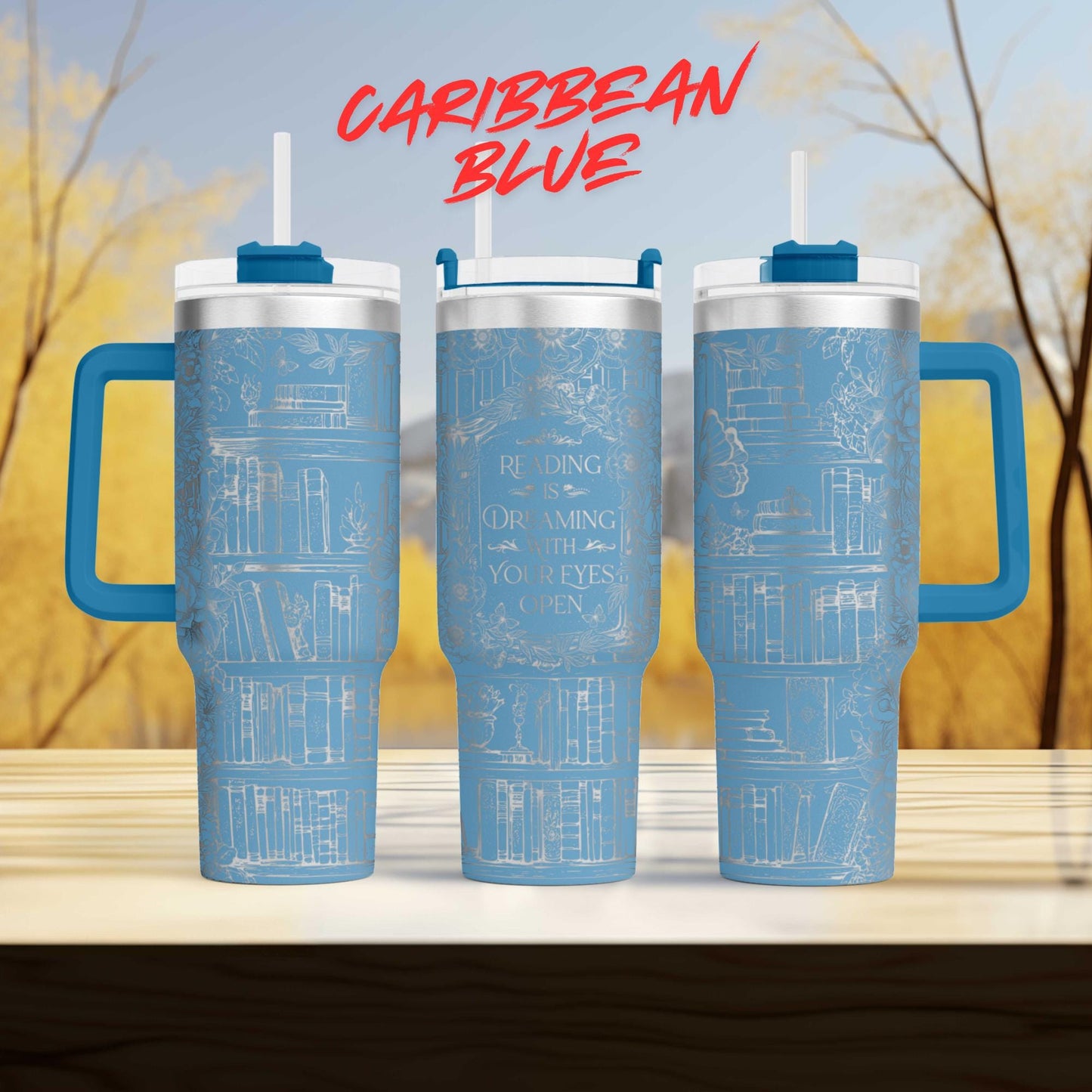 three blue tumblers with white text and illustrations on them, placed on a wooden surface against a blurred background of trees.