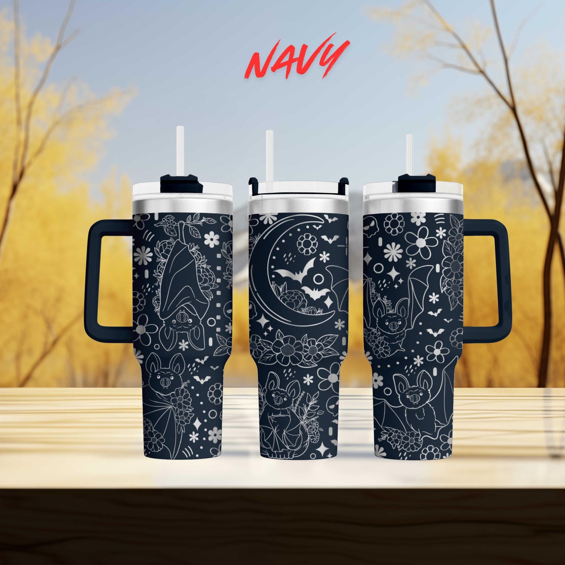 three black tumblers with white designs, including bats and flowers, against a blurred background of trees.