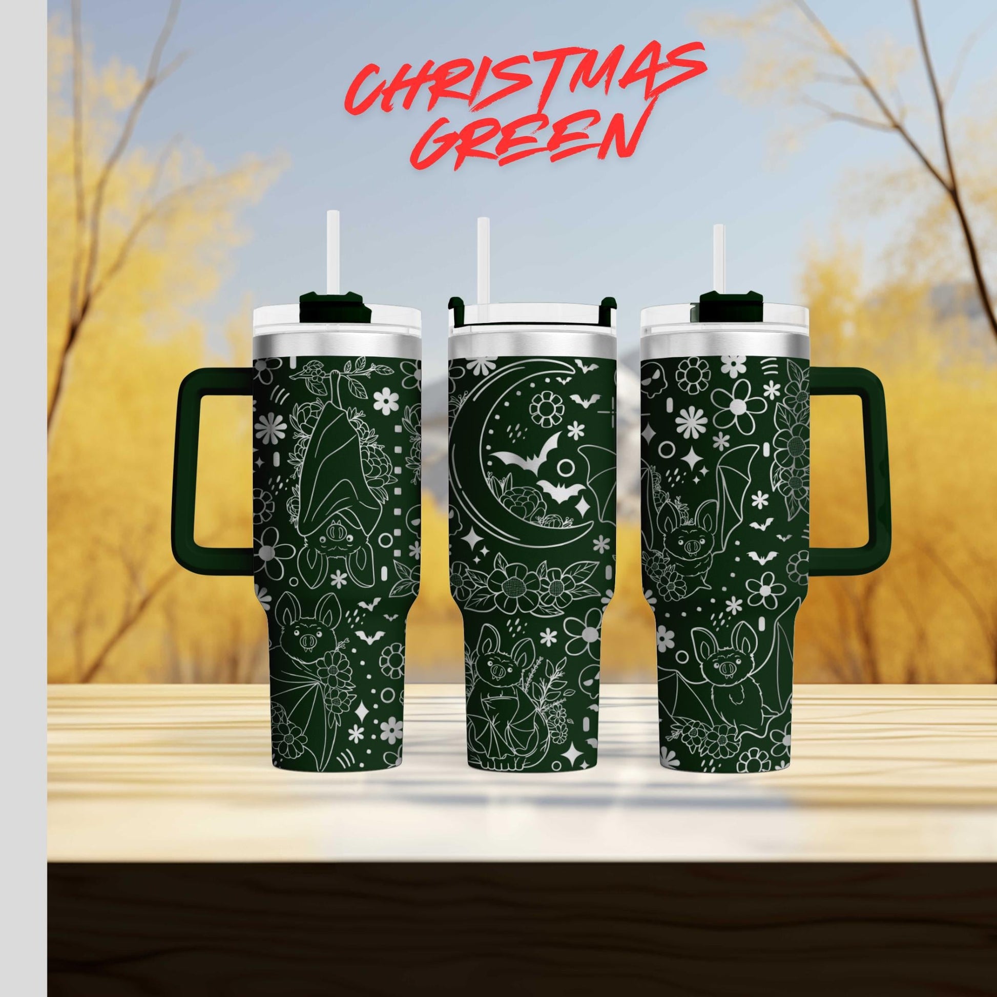 three green tumblers with white designs, including bats, flowers, and a crescent moon, against a blurred background of trees and a blue sky.