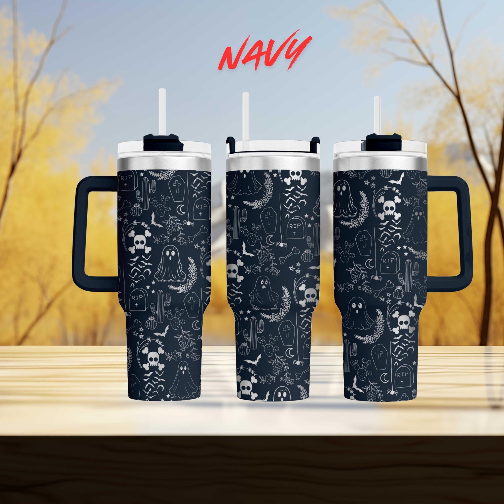 three black tumblers with a Halloween-themed design, including skulls, bats, and ghostly figures, displayed on a wooden surface against a blurred background of trees.