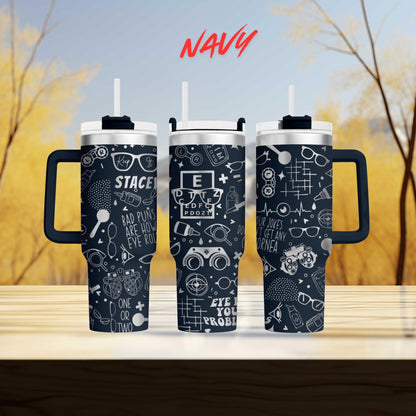 three black and white tumblers with various designs and text printed on them, placed on a wooden surface against a blurred background of trees.