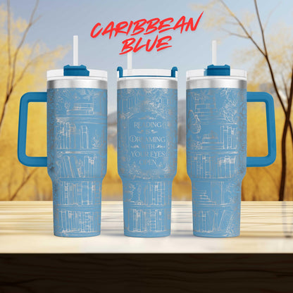 three blue tumblers with white text and illustrations on them, placed on a wooden surface against a blurred background of trees.
