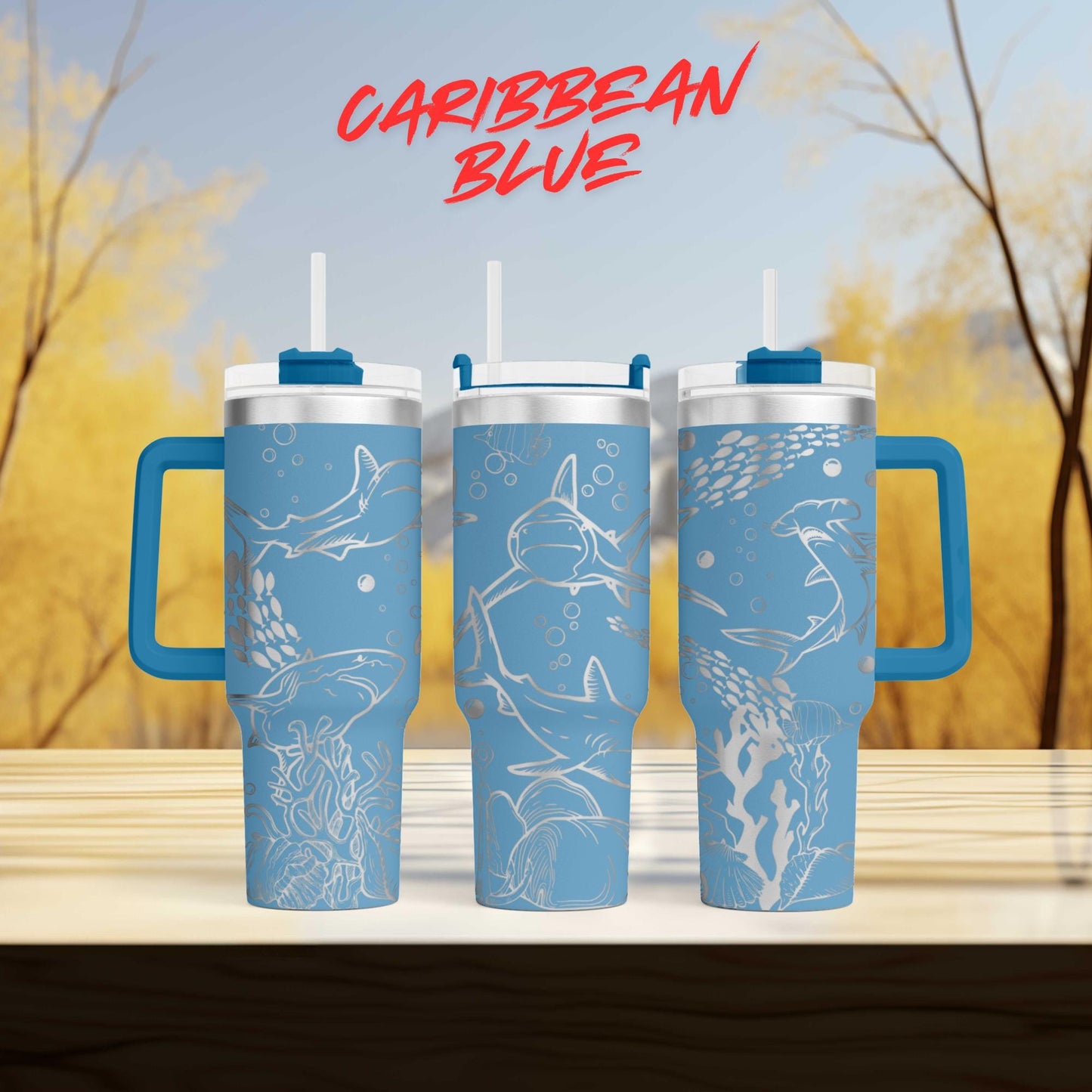 three blue tumblers with white designs, including sharks and bubbles, against a blurred background of trees.