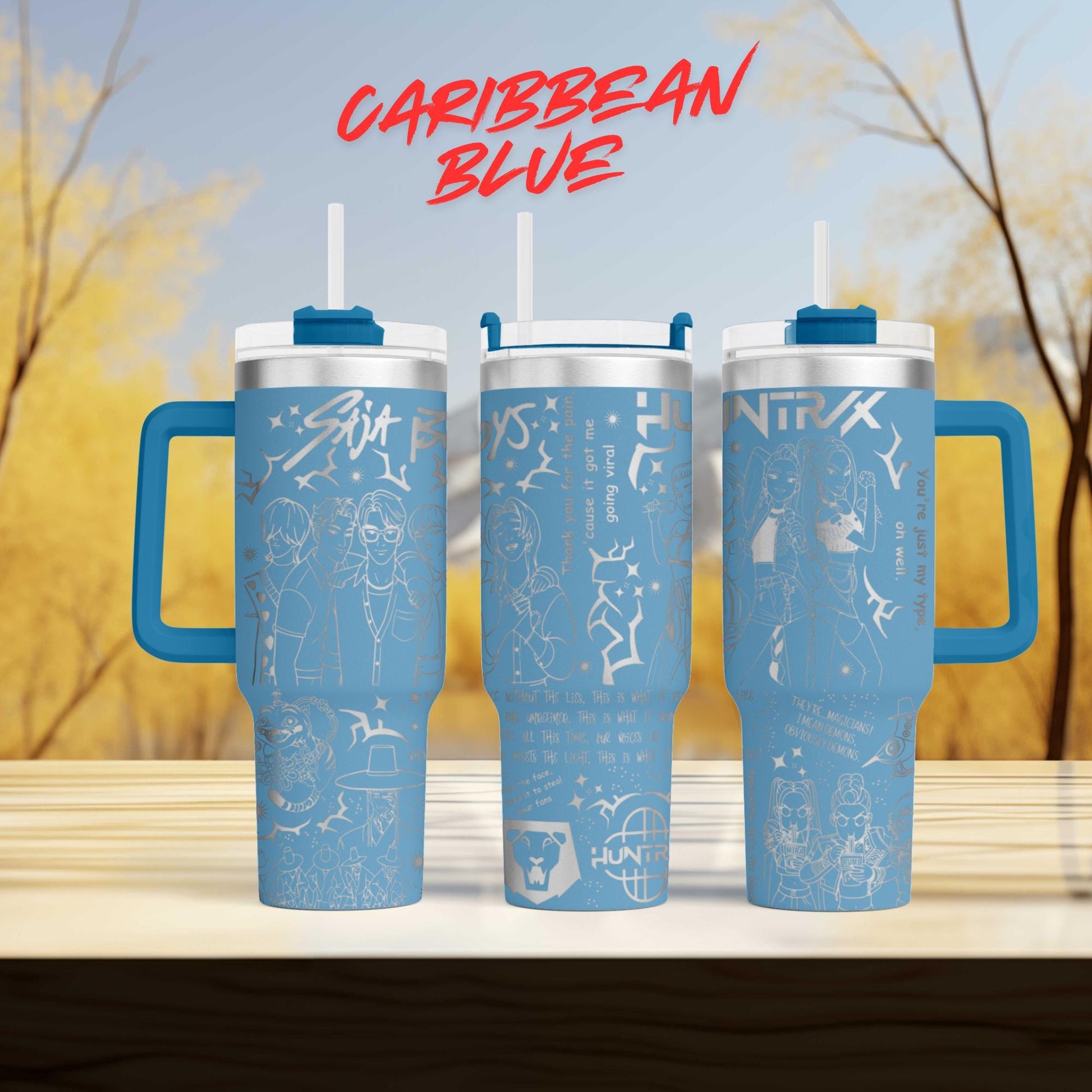 three blue tumblers with various designs and text on them, placed on a wooden surface against a blurred background of trees.