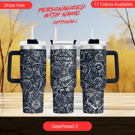 three black and white tumblers with a mechanical or industrial design, including gears, cogs, and other mechanical elements. The tumblers are labeled "Thomas" and "Gearshead 3", suggesting they are personalized with a name and model. The background of the image is blurred, but it appears to be a wooded area, possibly a forest or a park.