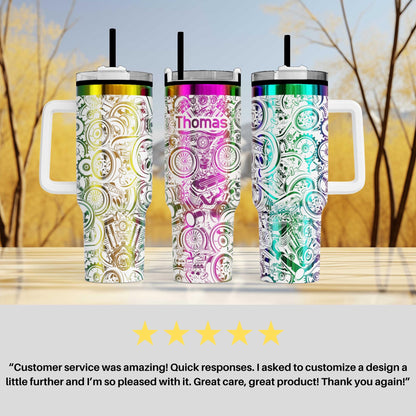 Three colorful, patterned insulated tumblers with handles, each featuring a unique design and color scheme. The tumblers are placed on a wooden surface against a blurred background of trees.